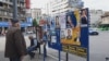 Panou cu afișe electorale în Piața Romană. Un bărbat privește promisiunile electorale care răzbat din mesajele candidaților. Lângă panou, proprietarul florăriei de vizavi stă pe un scaun, așteptând clienții.&nbsp;