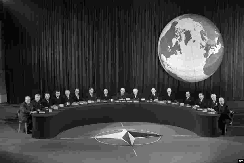 Конференция НАТО в Париже в 1957 году под эмблемой НАТО, принятой в 1953 году. Флаг НАТО тёмно-синего цвета — символ Атлантического океана. В центре изображена роза ветров в круге, от которой расходятся белые линии. Круг означает единство между членами альянса, а роза ветров — направления, в которые простирается путь к миру