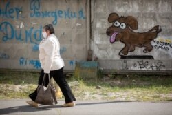 Жінка у медичній масці йде з торбами продуктів додому з магазину повз паркан зі смішним графіті