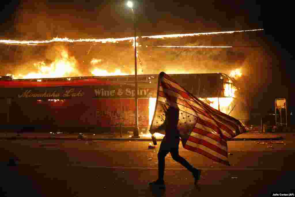Demonstrant sa američkom zastavom okrenutom naopako u znak protesta, hoda pored goruće zgrade 28. maja u Minneapolisu tokom protesta zbog smrti 46-godišnjeg Afroamerikanca Georgea Floyda. Floyd je preminuo 25. maja u bolnici. Na videu u kojem se prikazuje njegovo hapšenje vidi se bijeli policajac kako drži nogu na njegovom vratu dok Floyd govori da ne može disati.
